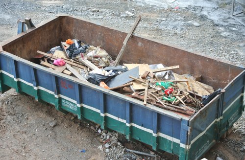 A large metal skip bin with a rusted orange interior, filled with mixed debris including wooden planks, broken pieces of drywall, plastic bags, and miscellaneous waste materials, borders a construction site with an uneven dirt surface and gravel in the background. The wooden planks vary in length and width, some appear weathered and splintered, while the plastic bags and packaging consist of black and transparent materials. The skip, positioned on a gravel and dirt ground, has a green and blue exterior with a corrugated metal texture, and appears to be ready for rubbish collection or removal. Natural daylight illuminates the scene, emphasizing the textured surfaces and contrasting colors of the waste and surrounding ground, reflecting typical site clearance activity that a professional rubbish removal service like Office Clearance Barnet might handle in the Barnet area.