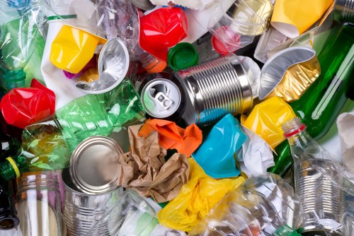 A close-up image of a large pile of mixed recyclable waste materials, including crushed aluminum cans, plastic bottles in various colours such as green, clear, and red, and crumpled cardboard, all scattered randomly on a flat surface. The aluminum cans feature ridged textures and metallic finishes, with some appearing slightly dented or squashed. Clear plastic bottles with visible caps are stacked and overlapping, while coloured plastic bottles and containers are dispersed throughout the pile. Wrinkled brown paper and crumpled supermarket shopping bags are also present among the plastic and metal debris. The environment suggests an indoor or outdoor waste collection point with natural light illuminating the scene, emphasizing the variety of textures and colours. This visual relates to rubbish removal services offered by Office Clearance Barnet for domestic or commercial clearance in the Barnet area, emphasizing the handling of mixed waste streams akin to those encountered during office or property clearances.