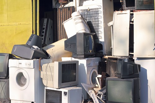 A collection of discarded household appliances and electronic waste piled together outdoors, including washing machines, microwaves, old computer monitors with screens and plastic casings, and other bulky electronic items, some leaning against each other and partially stacked. The appliances display a mix of materials such as metal, plastic, and glass, with variation in colours including white, black, and grey. The background features a metal shutter door and a wall, suggesting an industrial or storage area, possibly in Barnet or surrounding areas, often associated with waste removal services like those offered by Office Clearance Barnet. The scene appears to be in daylight, with natural light illuminating the objects, highlighting the textures of the metal and plastic surfaces. This image visually represents a typical scenario encountered during the collection and removal of electronic waste and bulky refuse, integral to rubbish clearance operations.