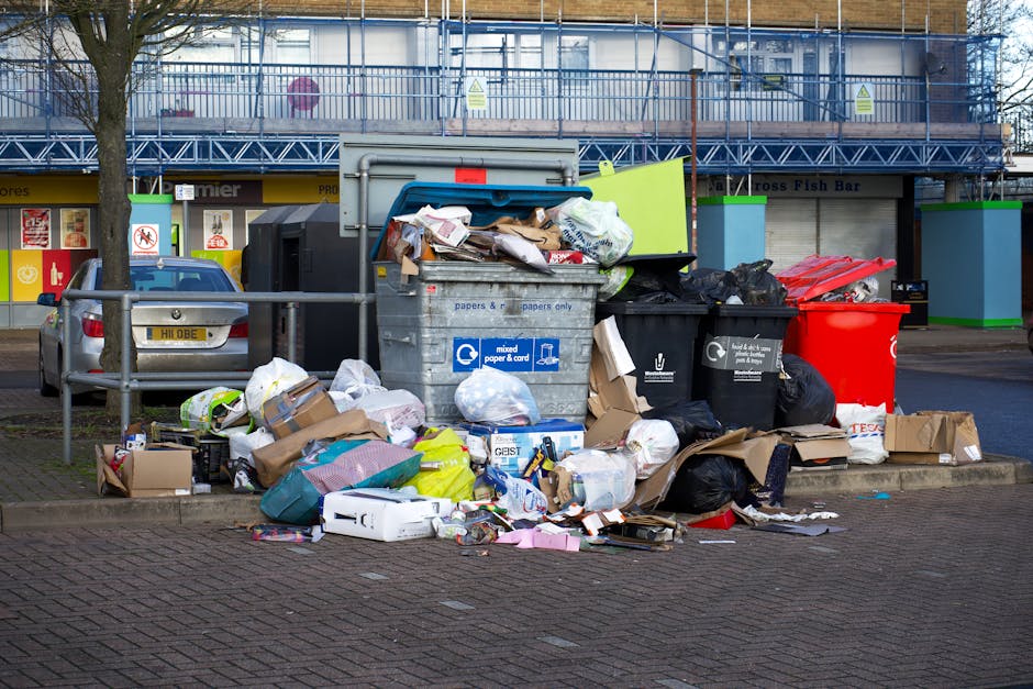A sanitation worker in a red and yellow uniform is operating a large red rubbish collection truck on a street, with the rear loading mechanism open and debris visible underneath. The worker is positio