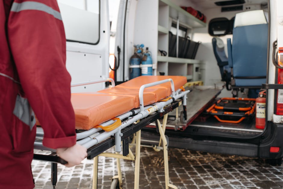Inside the rear of an emergency response vehicle, a person wearing a red uniform is partially visible, holding a wheeled stretcher with an orange padded mattress securely attached to its metal frame.