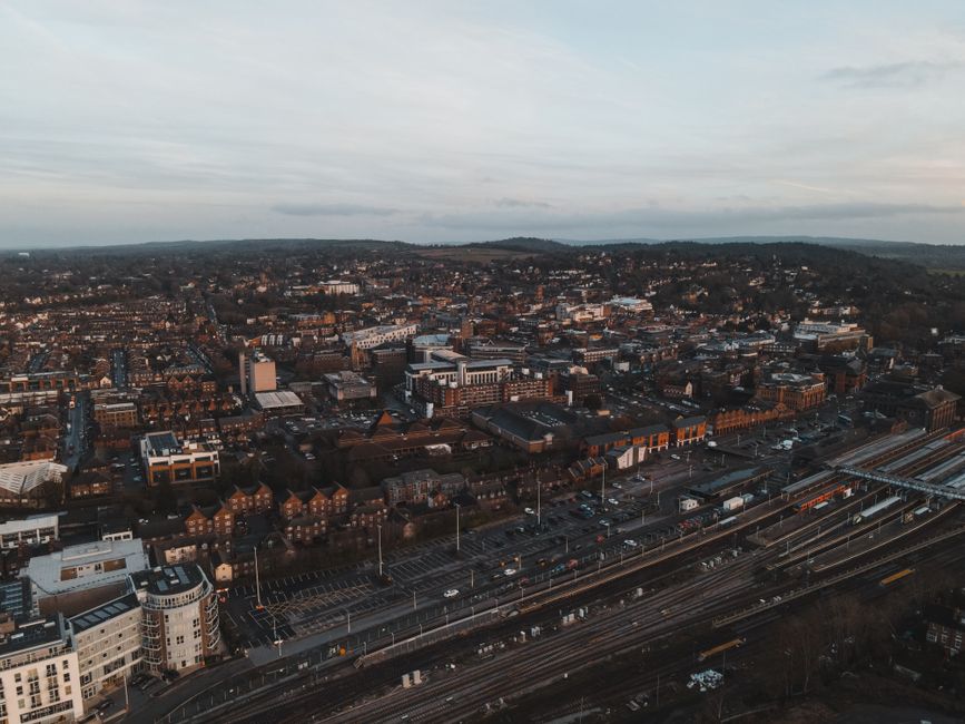 An aerial view of a densely built urban area showing a mixture of residential and commercial buildings in various architectural styles, with a prominent railway line running through the foreground. Th